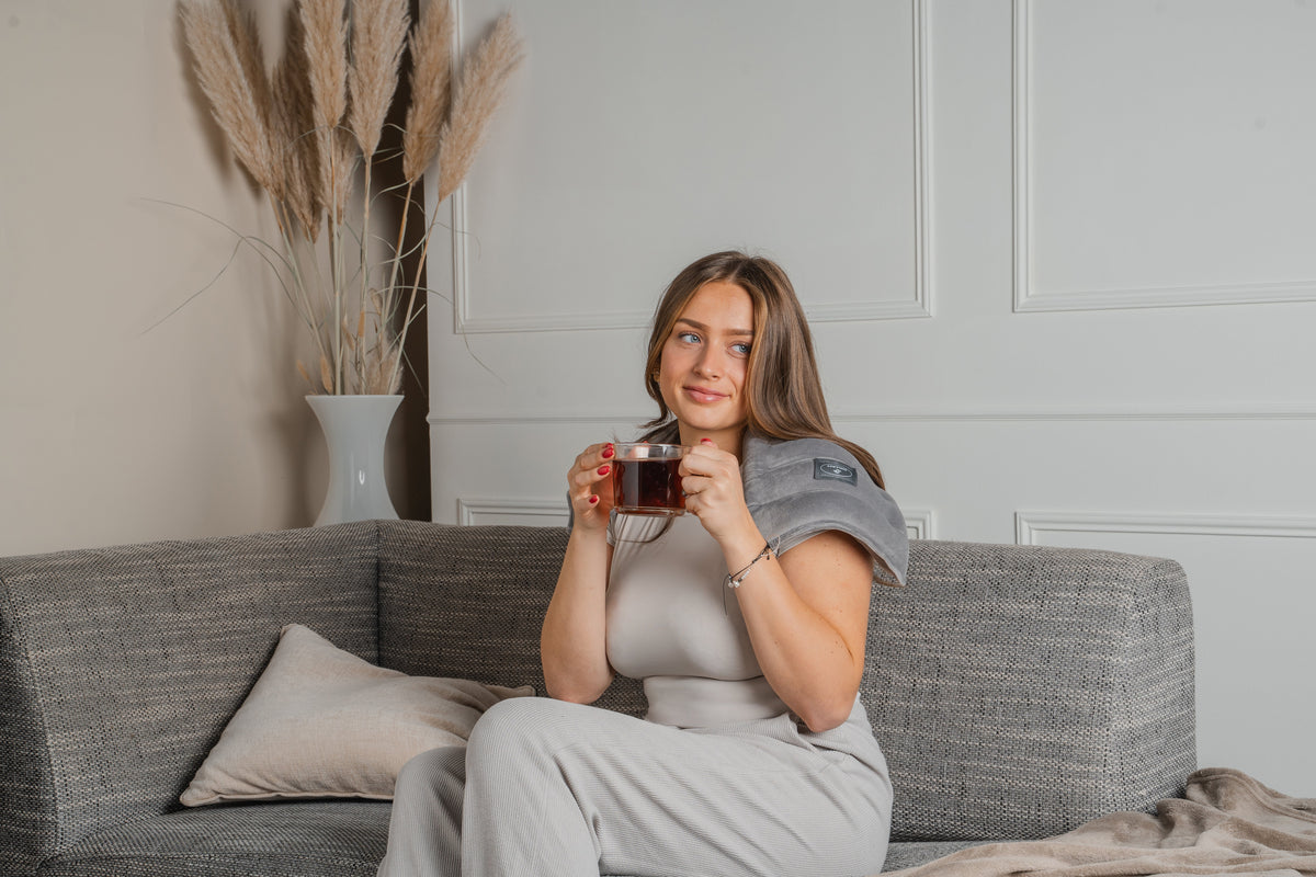 Vrouw op de bank met een warmtekussen op haar schouder en een kop thee